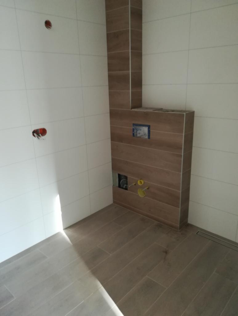 Wnętrze łazienki w trakcie prac wykończeniowych, widoczne białe płytki na ścianach, zabudowa stelaża WC wykończona płytkami imitującymi drewno ułożonymi poziomo, oraz ułożone podłogowe panele...