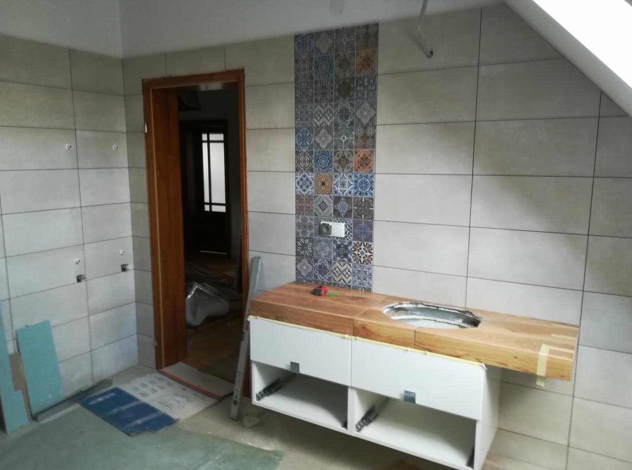Łazienka w trakcie prac wykończeniowych: ułożone płytki imitujące patchwork na ścianie, drewniany blat z otworem na umywalkę zamontowany na białej szafce, widoczne narzędzia i poziomica, w tle...