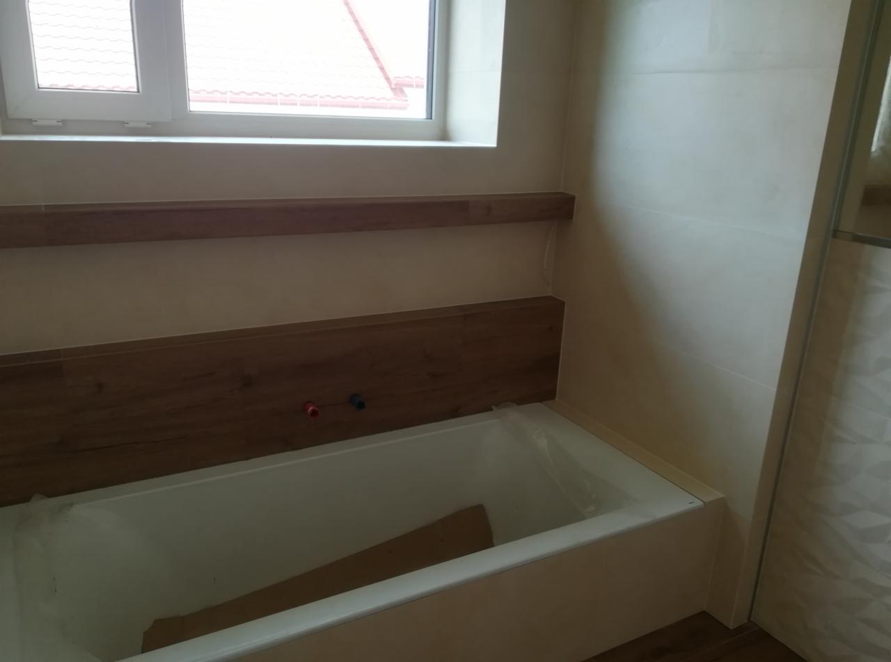 Wanna w trakcie obudowy, zabezpieczona folią, z widocznymi przyłączami wodnymi na tle beżowych płytek i drewnopodobnej okładziny.