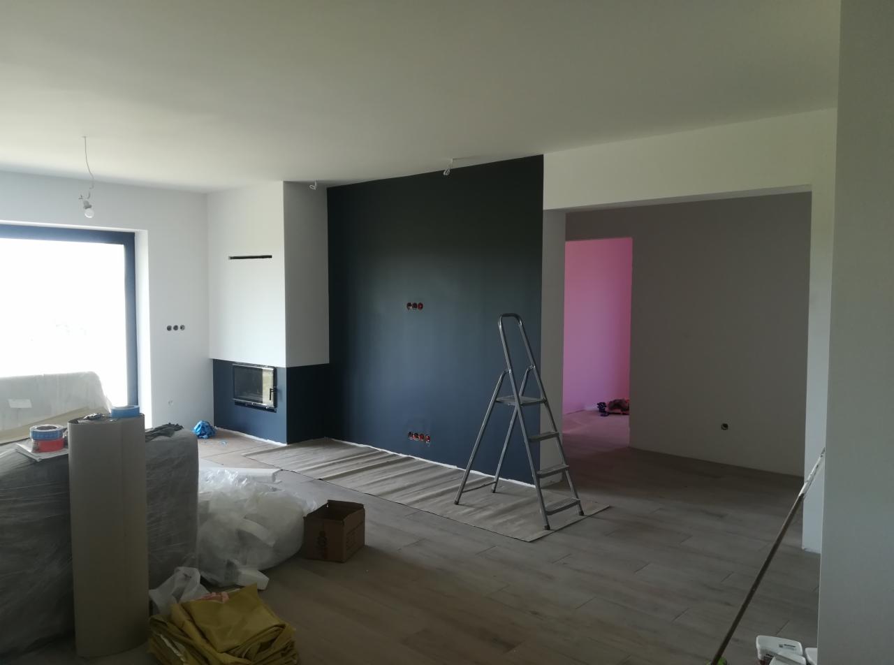 Pokój w trakcie malowania z ciemną ścianą, zabezpieczoną podłogą, drabiną i kominkiem w tle, widoczne gniazdka elektryczne.