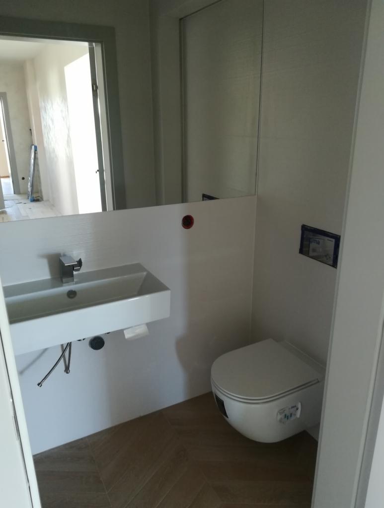 Nowoczesna łazienka z podwieszaną umywalką nablatową, toaletą w zabudowie i podłogą wyłożoną płytkami imitującymi drewno w jodełkę; widoczne elementy instalacji wodnej pod umywalką.