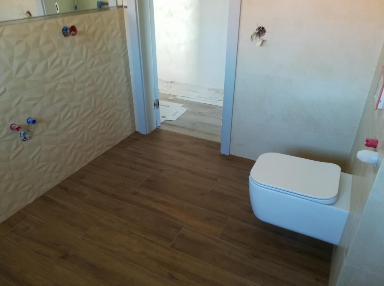 Nowoczesna łazienka w trakcie wykańczania: drewnopodobne płytki na podłodze, ściana z trójwymiarową fakturą, wisząca toaleta i widoczne instalacje hydrauliczne.