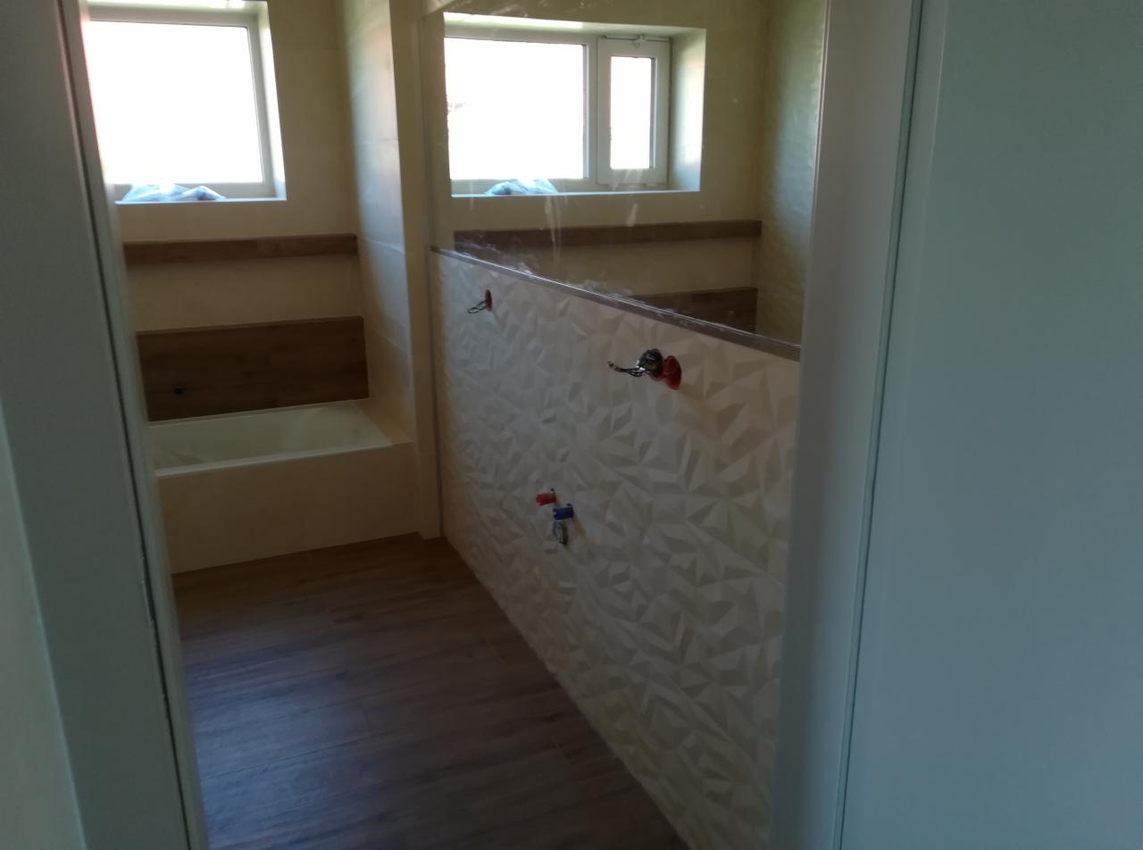 Wnętrze łazienki w trakcie prac wykończeniowych: ściana z trójwymiarową białą płytką, widoczne przyłącza wodne i fragment zabudowy wanny z drewnianym elementem dekoracyjnym, podłoga z paneli.