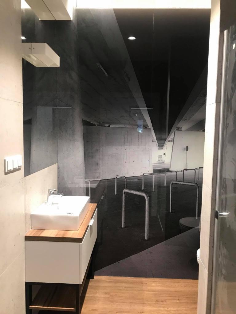 Szklana ściana z nadrukiem betonowego tunelu w łazience, widoczna umywalka z szafką i fragment toalety.