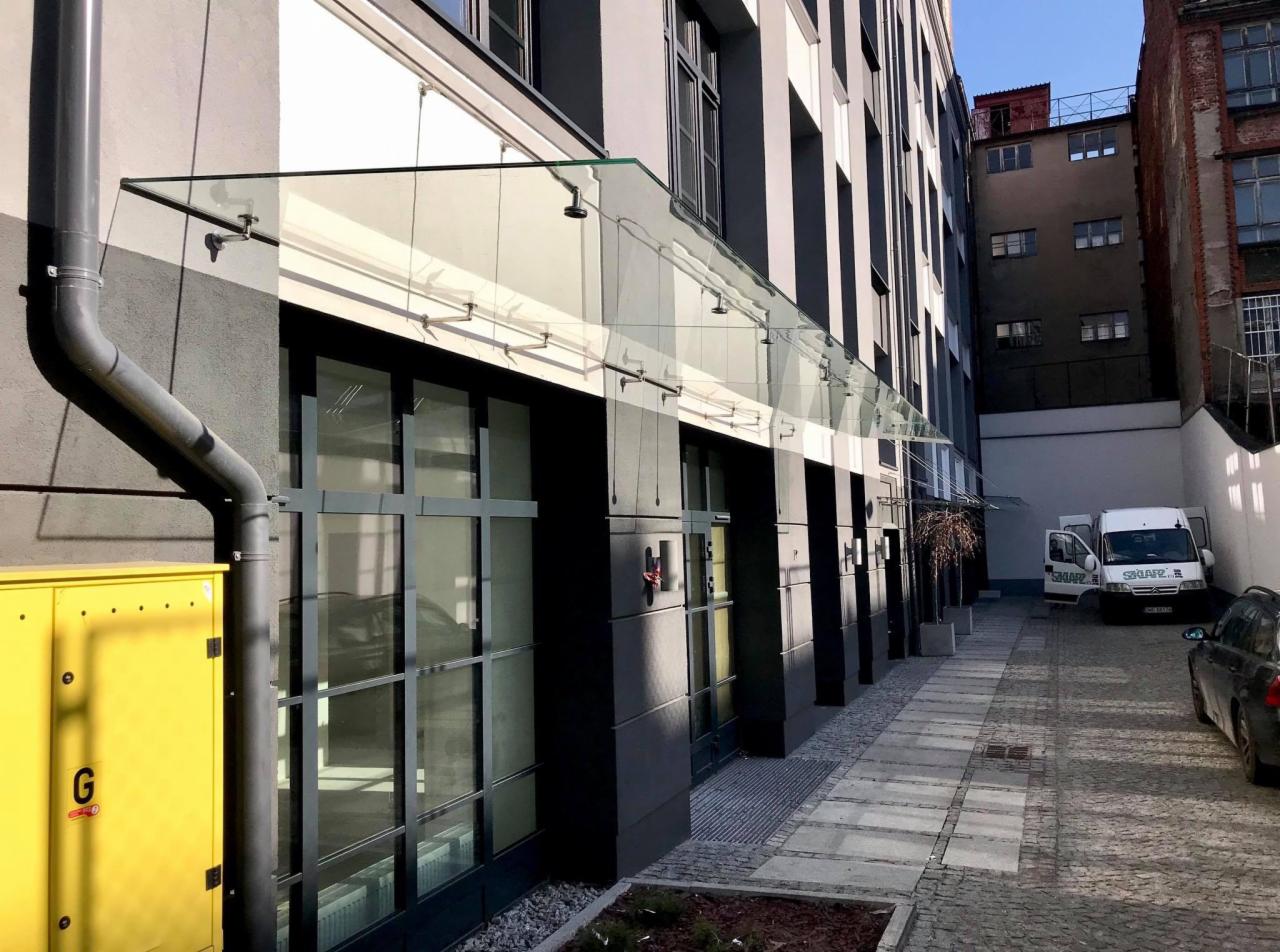 Szklane zadaszenie nad wejściem do nowoczesnego budynku biurowego z czarnymi ramami okiennymi i żółtą skrzynką elektryczną po lewej stronie, widok z poziomu chodnika.