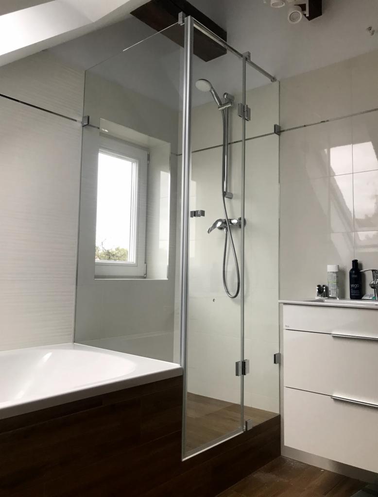 Szklana kabina prysznicowa z drzwiami otwieranymi na zewnątrz, zamontowana przy wannie, z widocznym oknem w tle i białą szafką z umywalką po prawej stronie.