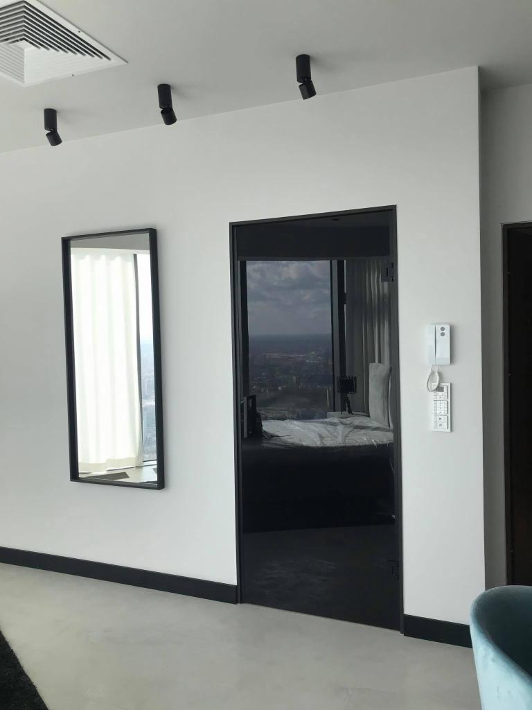 Nowoczesne wnętrze z lustrem w czarnej ramie na ścianie, obok drzwi z czarną ościeżnicą, widok na miasto w tle.