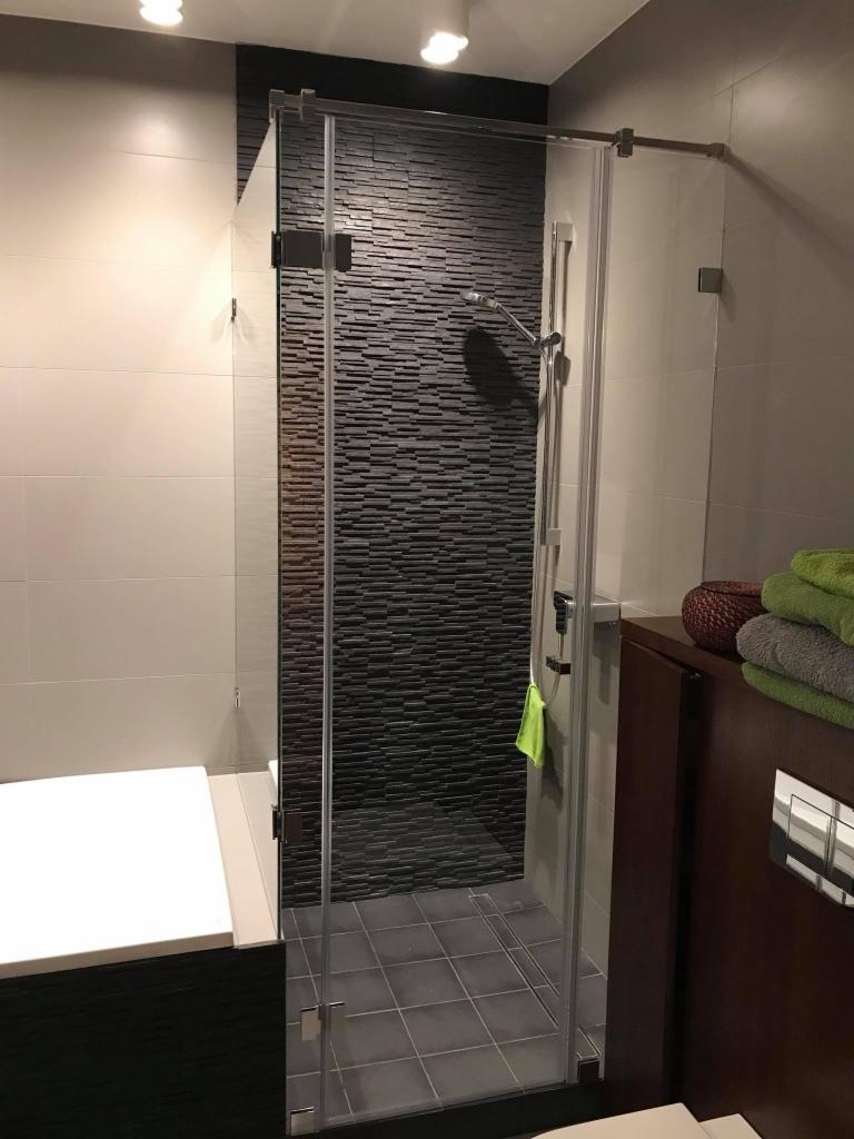 Szklana kabina prysznicowa z chromowanymi okuciami, przylegająca do wanny, z czarną ścianą wyłożoną wąskimi płytkami imitującymi kamień, widoczna zielona ściereczka na relingu.