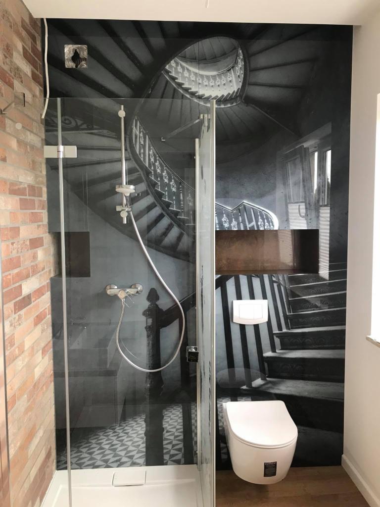 Szklana kabina prysznicowa z deszczownicą, obok toaleta i ściana z fototapetą przedstawiającą spiralne schody w czarno-białej tonacji, ceglana ściana po lewej stronie.