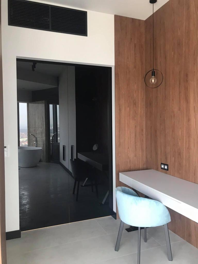 Czarne szklane drzwi przesuwne do łazienki z wolnostojącą wanną, widoczne z pokoju z drewnianą boazerią i toaletką z krzesłem w kolorze baby blue.