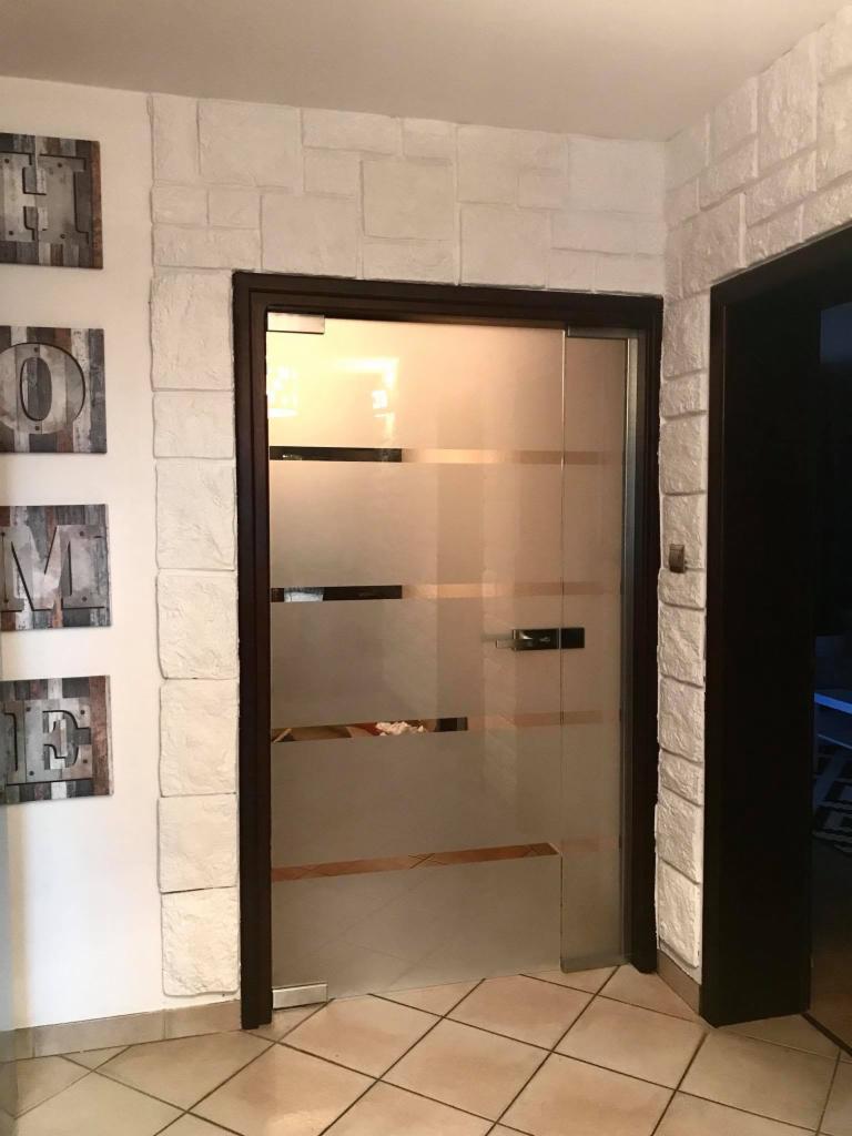 Szklane drzwi przesuwne z poziomymi pasami matowego szkła, w ciemnej ramie, zamontowane w ścianie wyłożonej dekoracyjną cegłą. Na ścianie obok wiszą drewniane litery HOME.