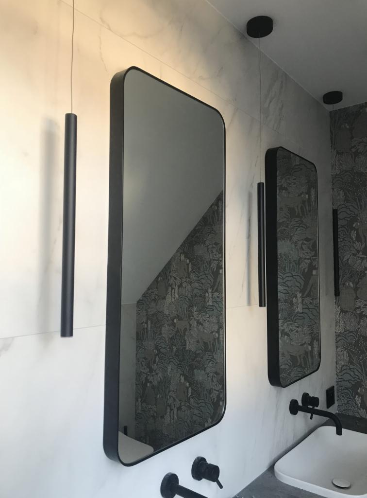 Dwa prostokątne lustra w czarnych ramach, zamontowane na ścianie z marmurowym wzorem, z widocznym odbiciem wzorzystej tapety w jednym z luster, czarne lampy tubowe wiszące obok luster, umywalka...