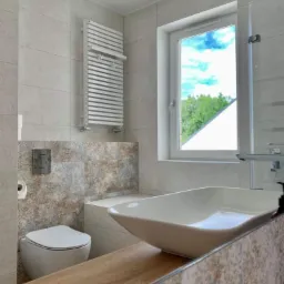 Nowoczesna łazienka z wiszącą toaletą, designerską umywalką nablatową i grzejnikiem drabinkowym, wykończona płytkami w odcieniach beżu i brązu, z widokiem na zieleń za oknem.