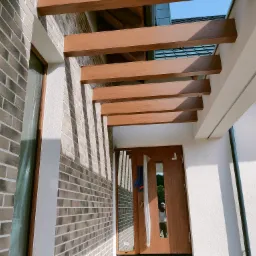 Nowoczesna elewacja domu z cegły klinkierowej w odcieniach szarości, z drewnianą pergolą nad wejściem. Widoczna faktura tynku i odbicie osoby w szybie drzwi.