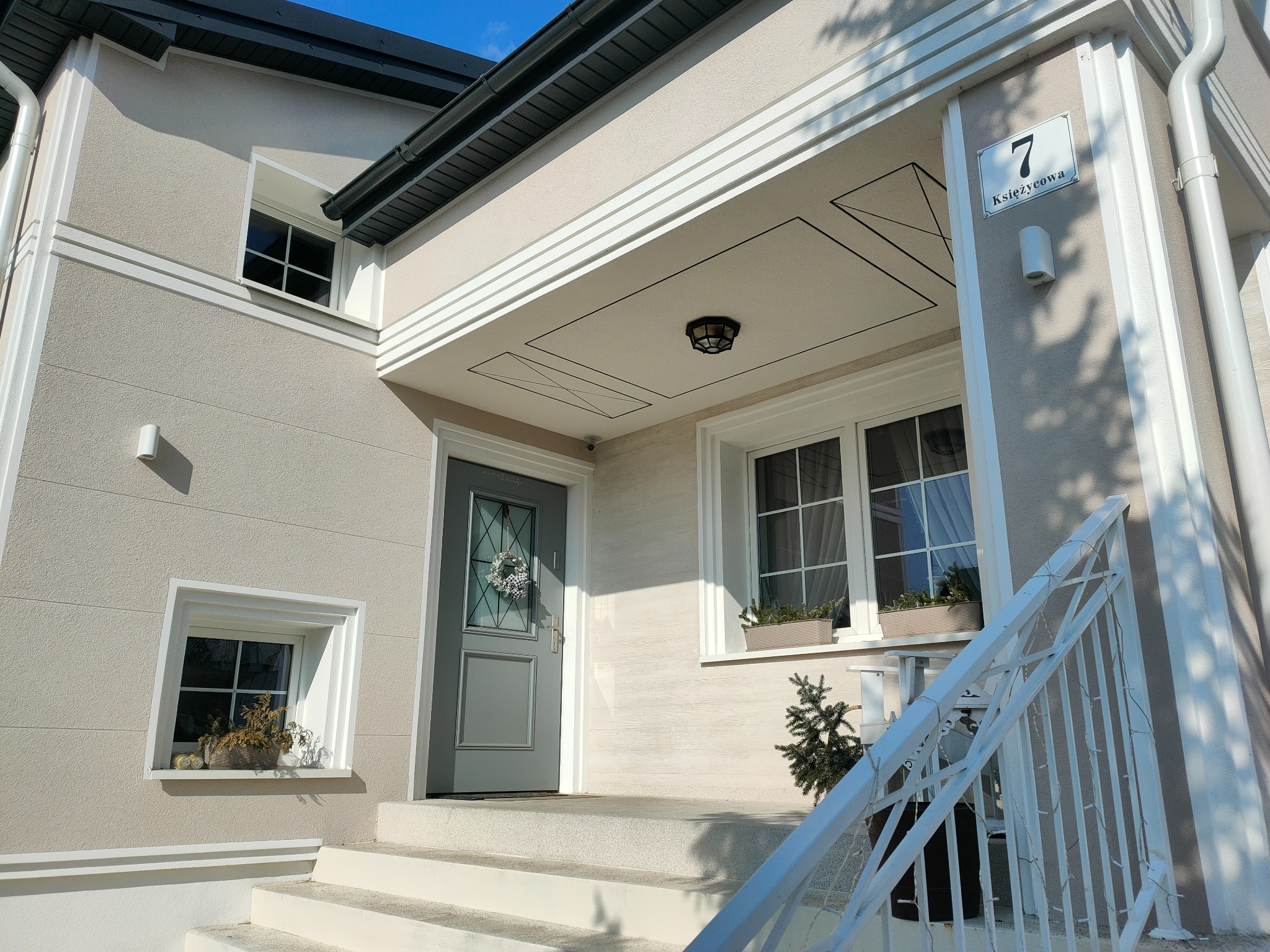 Jasna elewacja domu z dekoracyjnymi listwami, numerem domu '7 Księżycowa', szarą stolarką okienną i drzwiową oraz schodami z białą balustradą.