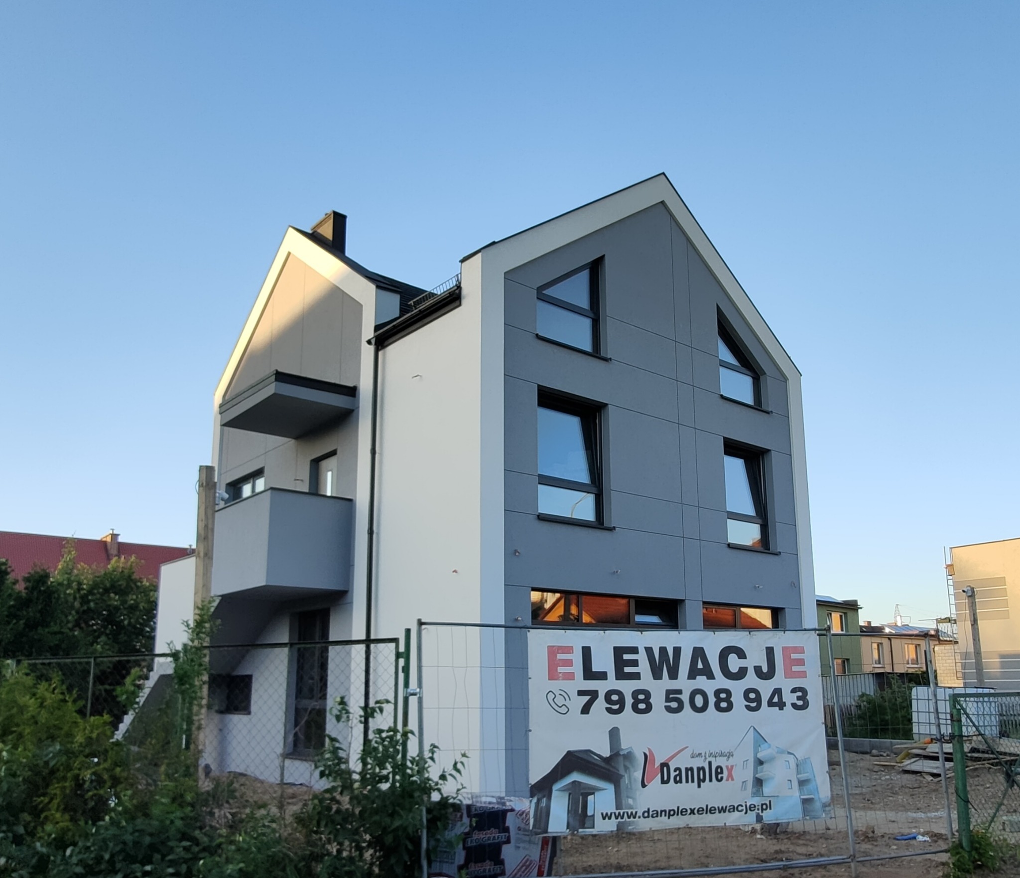 Nowoczesny dom z szarą elewacją panelową i białymi akcentami, widoczny przez ogrodzenie budowlane z reklamą firmy elewacyjnej Danplex