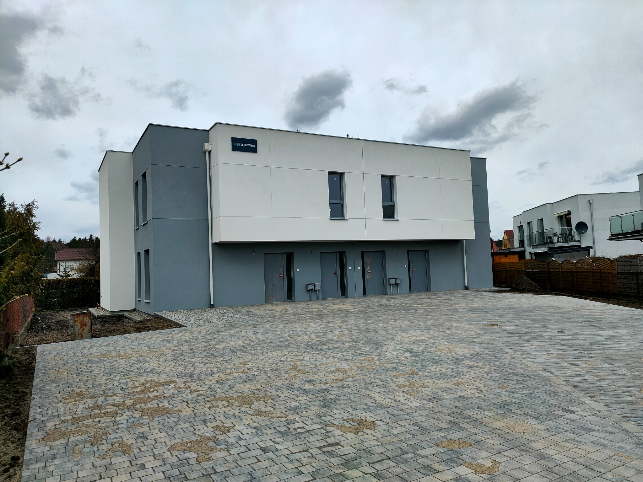 Dwukondygnacyjny budynek z szarą i białą elewacją, widoczny z poziomu brukowanego podjazdu, z logo firmy budowlanej na górze.