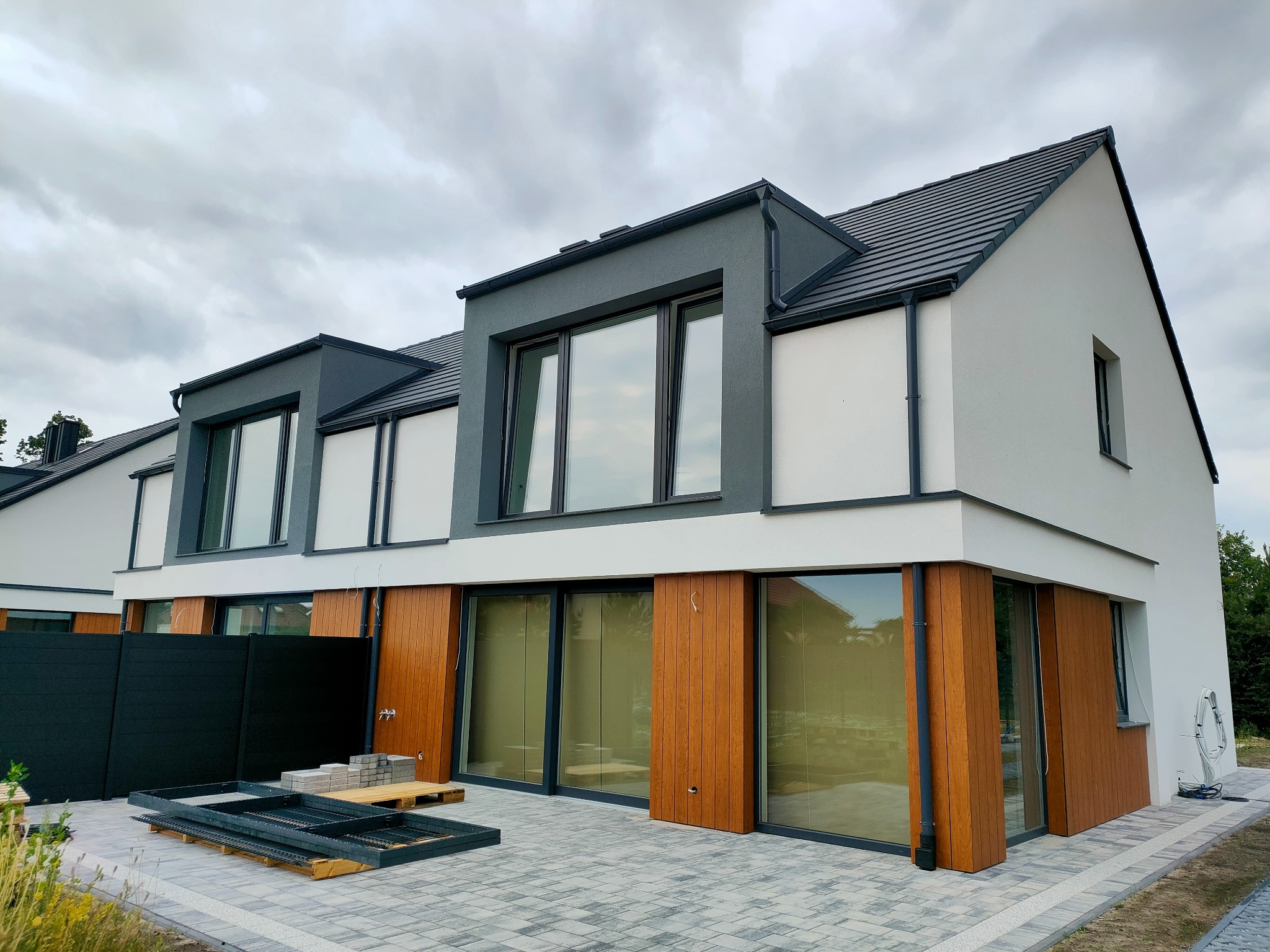 Nowoczesny budynek jednorodzinny z szarą i białą elewacją, drewnianymi panelami, dużymi oknami i kostką brukową na podjeździe, widoczne elementy konstrukcyjne i materiały budowlane.
