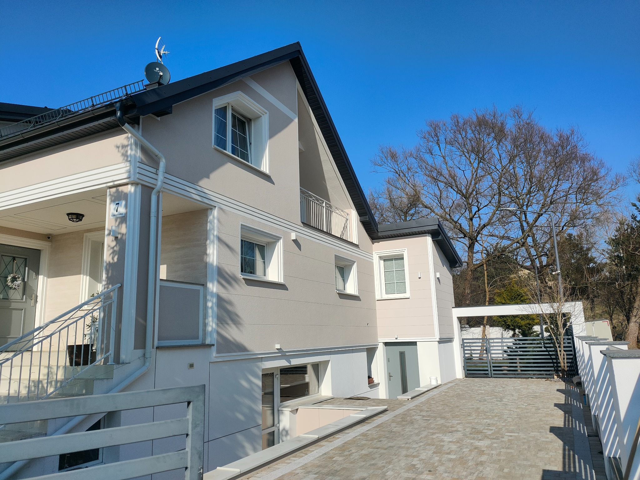 Dwupiętrowy dom z beżową elewacją, białymi obramowaniami okien i balkonem, widoczny z poziomu brukowanego podjazdu w słoneczny dzień.