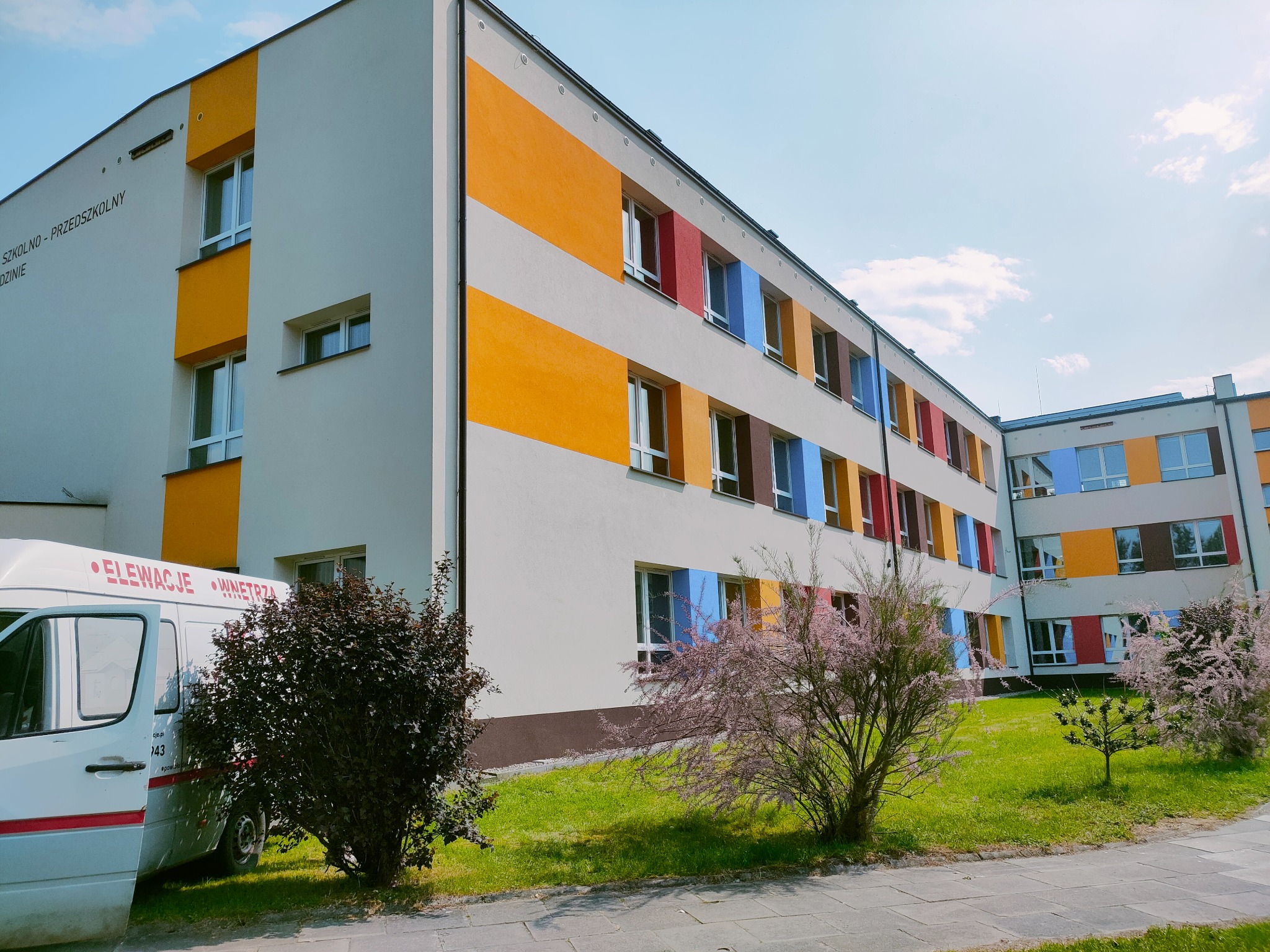 Odnowiona elewacja budynku szkolnego z kolorowymi pasami i obramowaniami okien, widoczny firmowy bus z napisem 'Elewacje Wnętrza'.