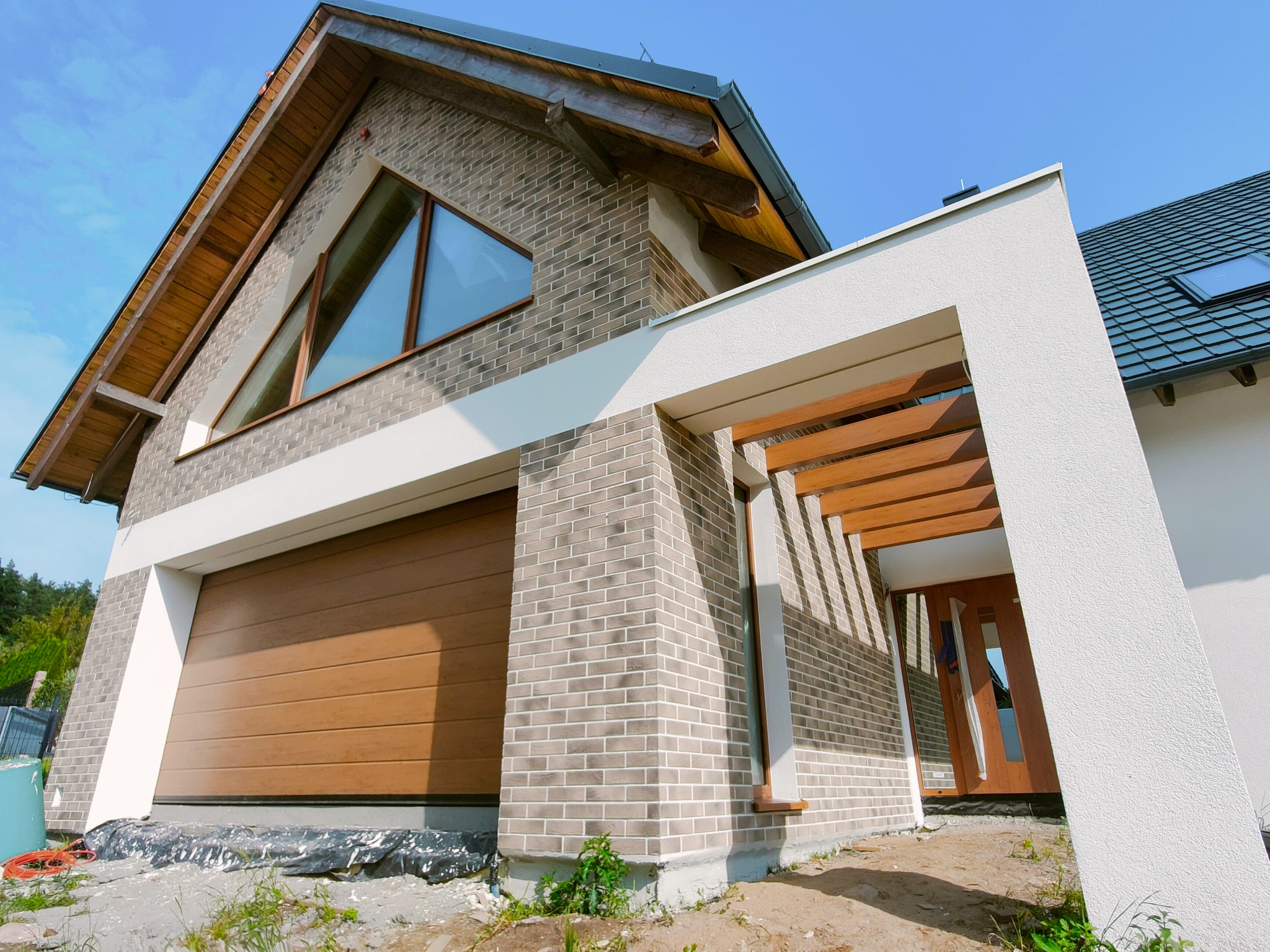 Nowoczesny dom z elewacją z cegły klinkierowej i drewnianymi elementami wykończeniowymi, garaż z bramą segmentową, błękitne niebo w tle.