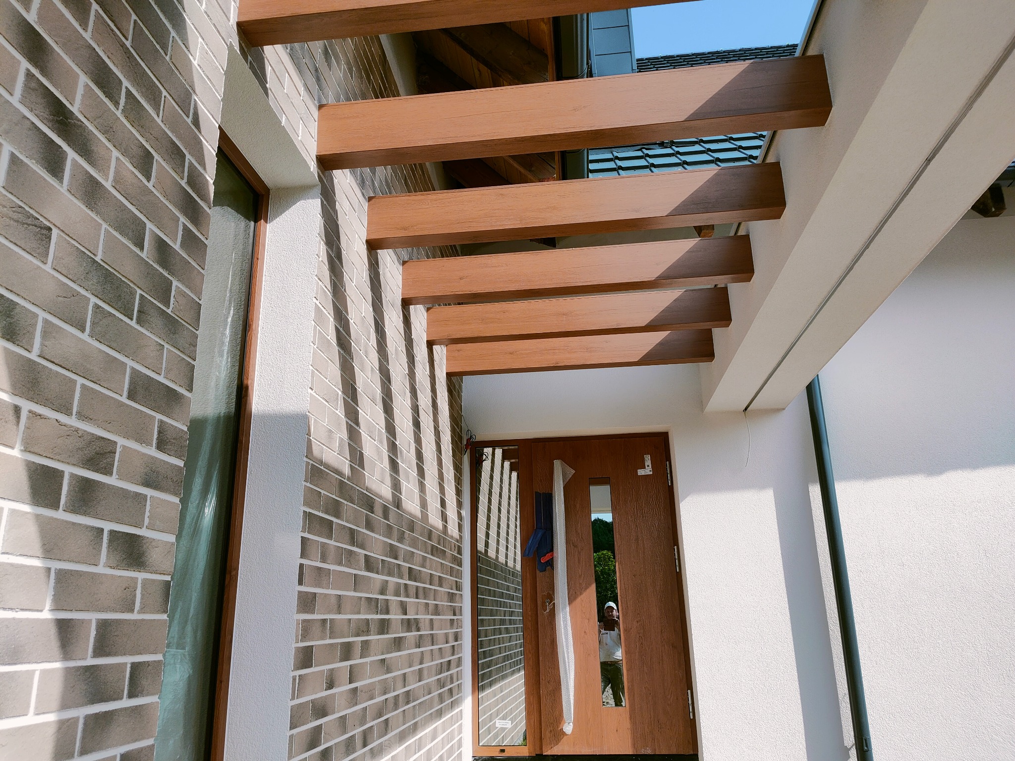 Nowoczesna elewacja domu z cegły klinkierowej w odcieniach szarości, z drewnianą pergolą nad wejściem. Widoczna faktura tynku i odbicie osoby w szybie drzwi.