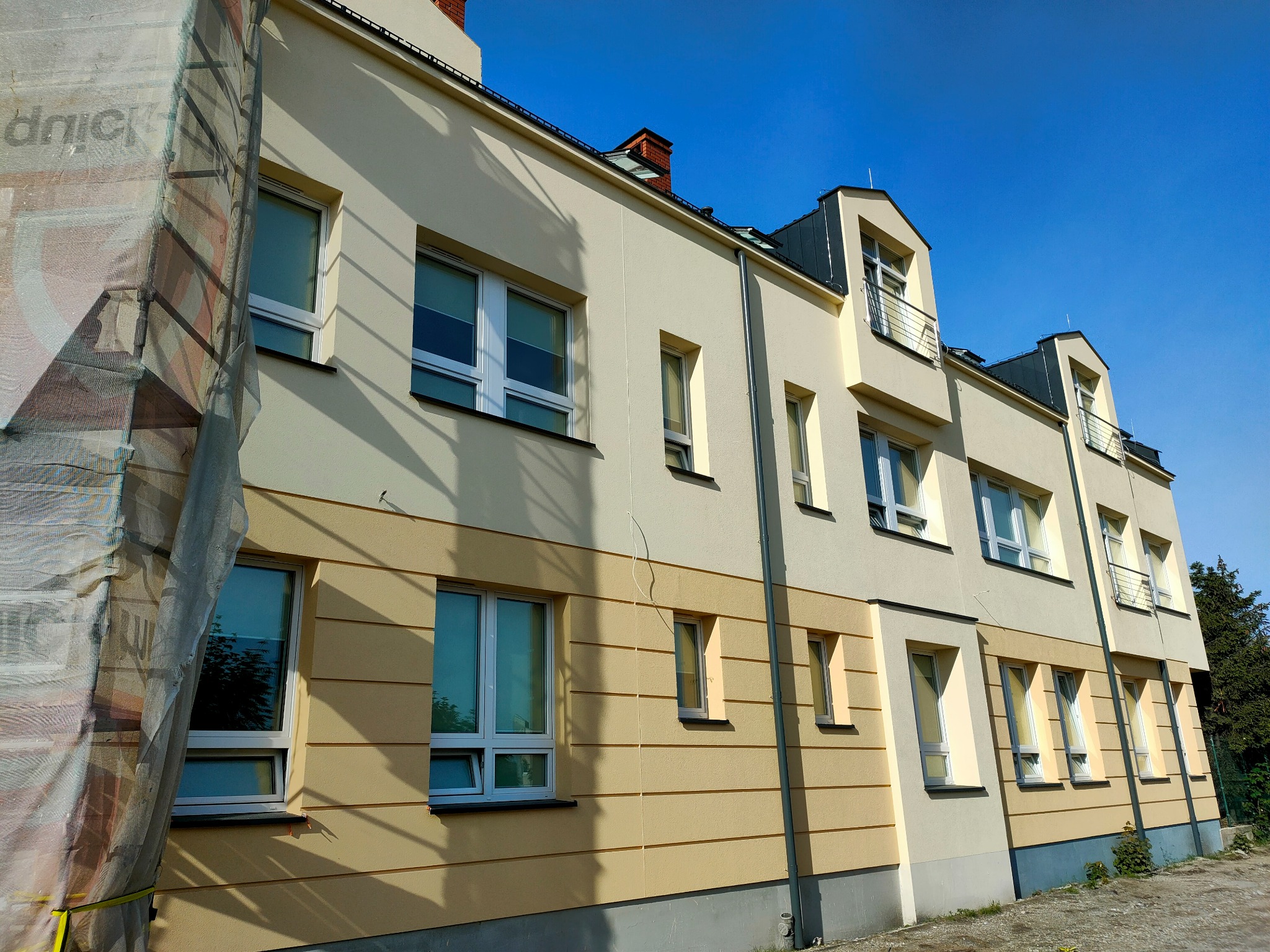 Dwukolorowa elewacja budynku wielorodzinnego w odcieniach beżu i żółci, częściowo osłonięta siatką rusztowania. Widoczne okna i balkony, błękitne niebo w tle.