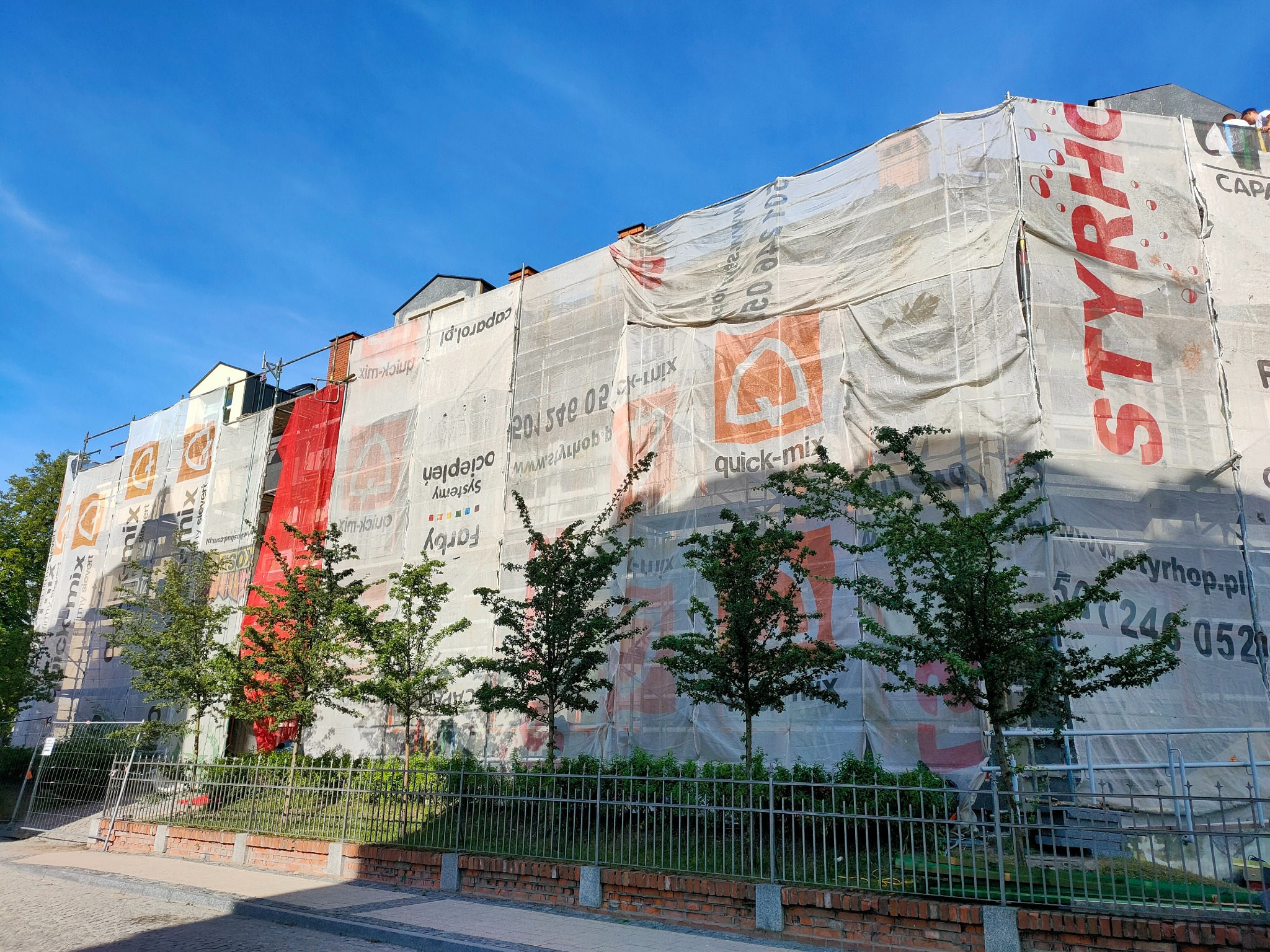 Budynek w trakcie prac elewacyjnych, osłonięty siatką z reklamami. Widoczne rusztowania, drzewa przed budynkiem i fragment niebieskiego nieba.