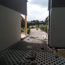 Niedokończona ścieżka z ażurowych betonowych płyt, widok z poziomu gruntu pomiędzy dwoma budynkami, z narzędziami i stertą płyt w tle.