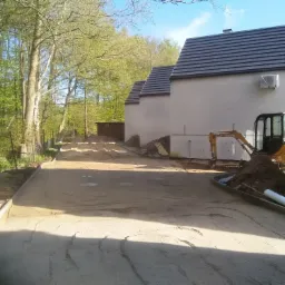 Przygotowanie podłoża pod nawierzchnię, widoczny mały traktor Case, obrzeża betonowe, budynki w stanie surowym, teren budowy.