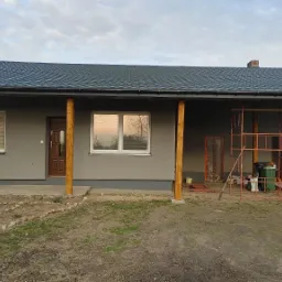 Dom jednorodzinny z szarą elewacją, drewnianymi kolumnami podtrzymującymi dach, widoczne rusztowanie i teren przed domem z ułożonymi kamieniami.