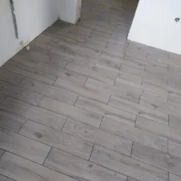 Ułożone, jasne płytki podłogowe imitujące drewno w nowym pomieszczeniu z surowymi ścianami i wystającymi instalacjami.