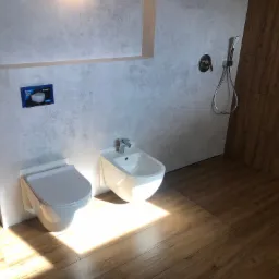 Nowoczesna łazienka w Pustej Kuźnicy z podwieszaną toaletą i bidetem, wykończona płytkami imitującymi beton i drewnem na podłodze oraz ścianie kabiny prysznicowej.