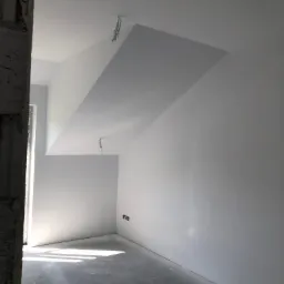 Poddasze po gładziowaniu, widoczne świeże, białe ściany i sufit ze skosem, surowy beton na podłodze z plamami farby, światło dzienne wpadające przez okno, niedokończona kolumna z cegieł po lewej...