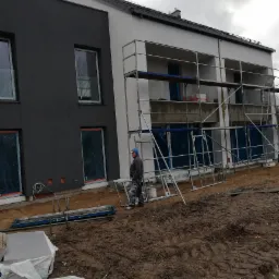 Dwukondygnacyjny budynek w trakcie prac elewacyjnych, z szarym i białym tynkiem, rusztowaniami, zabezpieczonymi oknami i pracownikiem w ubraniu roboczym na błotnistym terenie.