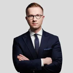 Portret młodego mężczyzny w okularach i granatowym garniturze, z rękami skrzyżowanymi na piersiach, na jednolitym szarym tle.