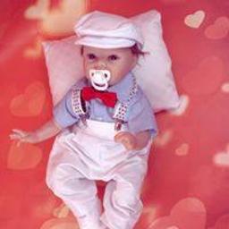 AnDZIA Ubranka Tworzone z Pasją - Lalka niemowlęca w białym ubranku, kapeluszu i z czerwoną muszką leżąca na poduszce na tle w serduszka.