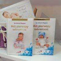 Dwa identyczne albumy pamiątkowe 'Mój pierwszy sakrament' z różnymi zdjęciami niemowląt na okładkach, ustawione obok siebie na białej półce, w tle koszyk z wikliny i pudełko z napisem 'Zaproszenie...
