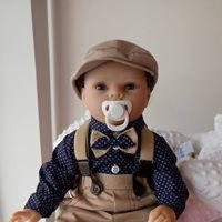 Lalka reborn w eleganckim stroju dziecięcym: koszula w granatowe kropki z muszką, beżowe szelki i spodnie, kaszkiet na głowie, smoczek w ustach.