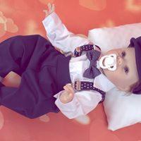 Uroczy niemowlak w eleganckim, granatowym garniturku z muchą w kropki, leżący na białej poduszce z smoczkiem w ustach, na tle w odcieniach czerwieni z rozmyciem w kształcie serc.