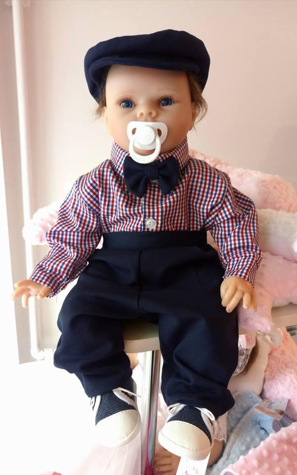 Ubrana lalka w elegancki, granatowy strój z muchą i kaszkietem, siedząca na krześle, w tle pluszowe zabawki.