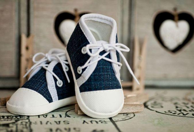 Ręcznie szyte, miniaturowe, dżinsowe trampki dla niemowląt z białymi sznurówkami na drewnianym podeście, dekorowane sercami i klamerkami w tle.