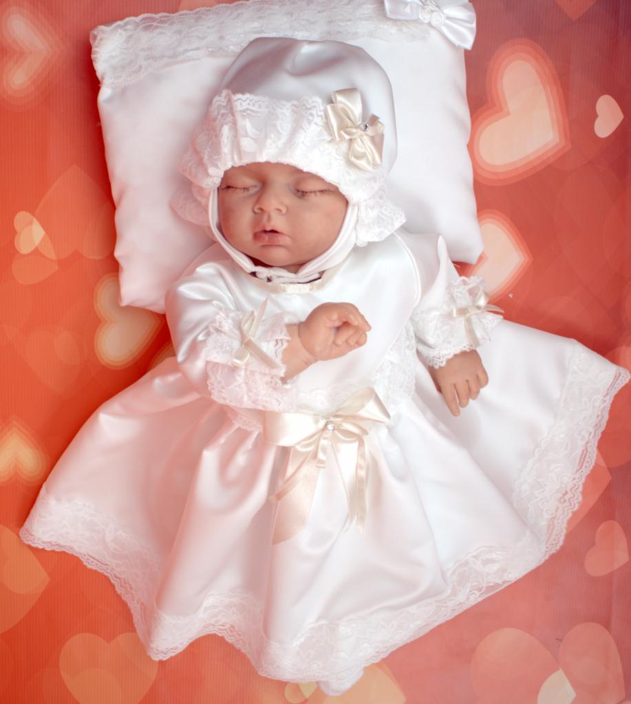 Śpiące niemowlę w białej, satynowej sukience i czepku z koronką, leżące na poduszce na tle w serduszka.