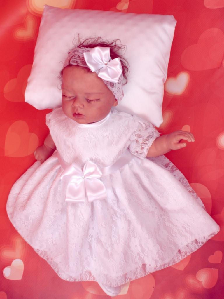 Śpiąca lalka w koronkowej sukience z kokardą, leżąca na białej poduszce na tle w serduszka, zdjęcie wykonane w Sochaczewie.