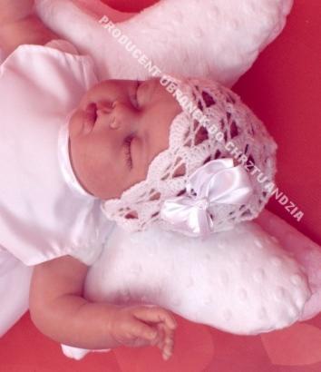 Śpiące niemowlę w białej, koronkowej czapeczce z satynową kokardką, ubrane w białą sukienkę, leżące na białej poduszce w kształcie gwiazdy na czerwonym tle.