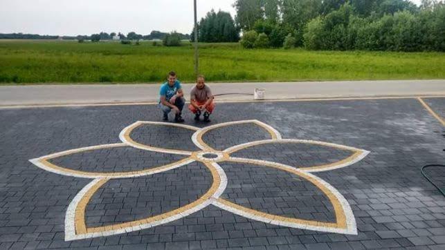 Dwóch mężczyzn przykuca na parkingu z kostki brukowej z wzorem kwiatowym w kolorach szarym, żółtym i białym; w tle widok na zielone pole i drzewa.