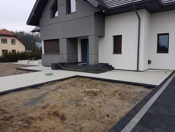 Świeżo ułożony jasnoszary chodnik z obramowaniem z ciemnej kostki wokół domu z białymi ścianami i szarym dachem, widoczny fragment trawnika przygotowany pod nasadzenia, zwinięty zielony kabel...