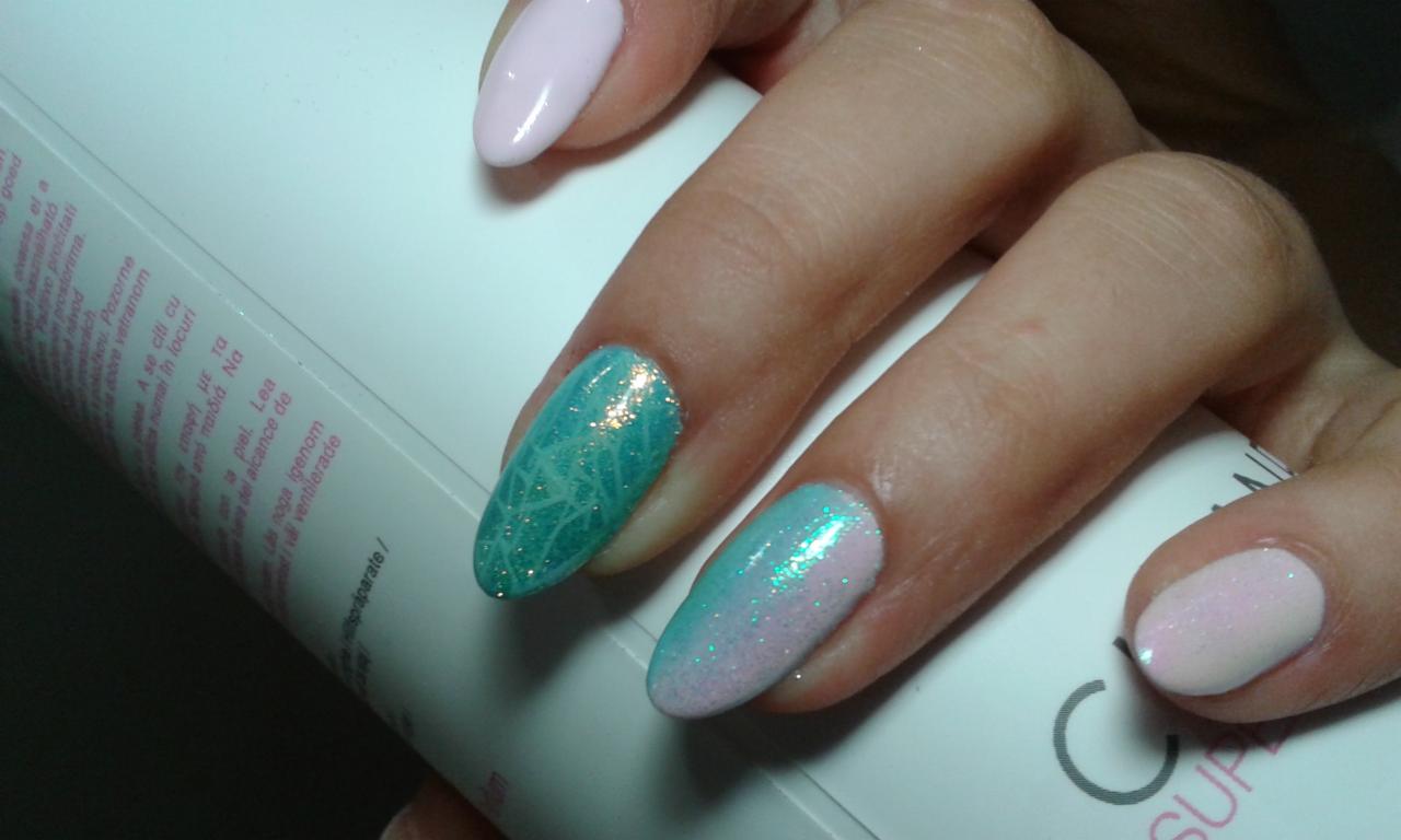 Dłonie z pomalowanymi paznokciami: dwa jasnoróżowe, jeden turkusowy z motywem pajęczyny i brokatem, jeden z przejściem kolorów od różu do turkusu z brokatem, na tle białej butelki z napisami.