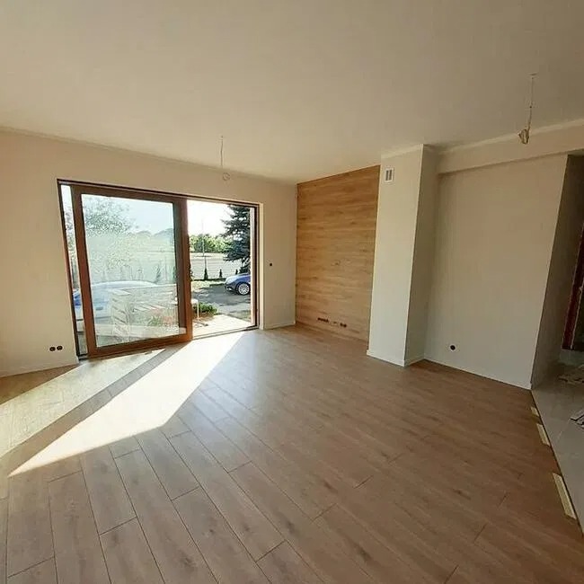 Jasne, przestronne wnętrze z drewnianą podłogą i akcentem z paneli drewnopodobnych na ścianie, widok na zewnątrz przez duże, przesuwne okno.