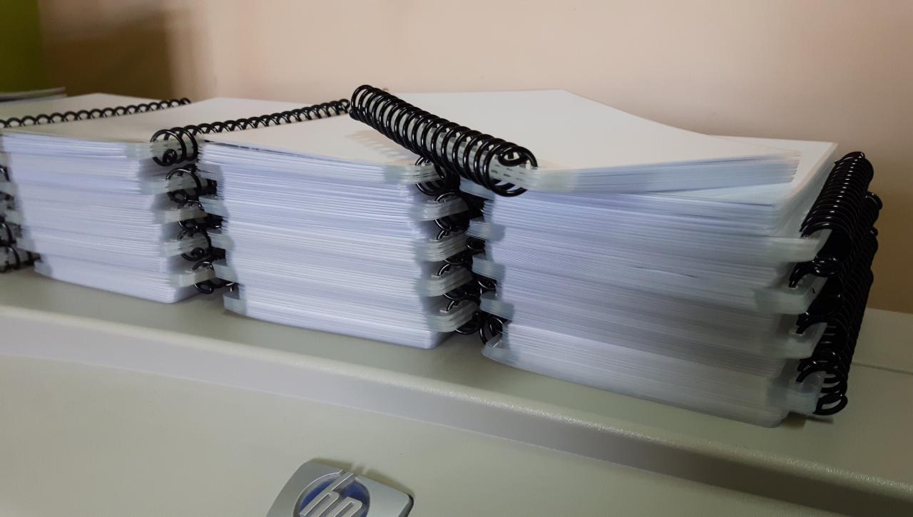 Stosy świeżo wydrukowanych, zbindowanych notatników z czarną spiralą, leżące na drukarce HP, gotowe do użycia.